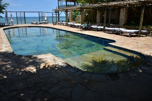 A clear swimming pool with lounge chairs and umbrellas, surrounded by stone flooring and a wooden pergola.