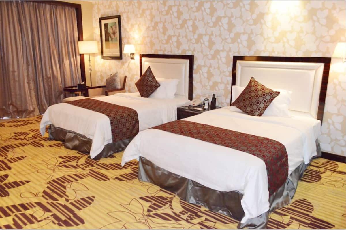 A hotel room with two beds, each with a white bedspread and a patterned throw blanket. There are bedside tables with lamps and a painting on the wall.