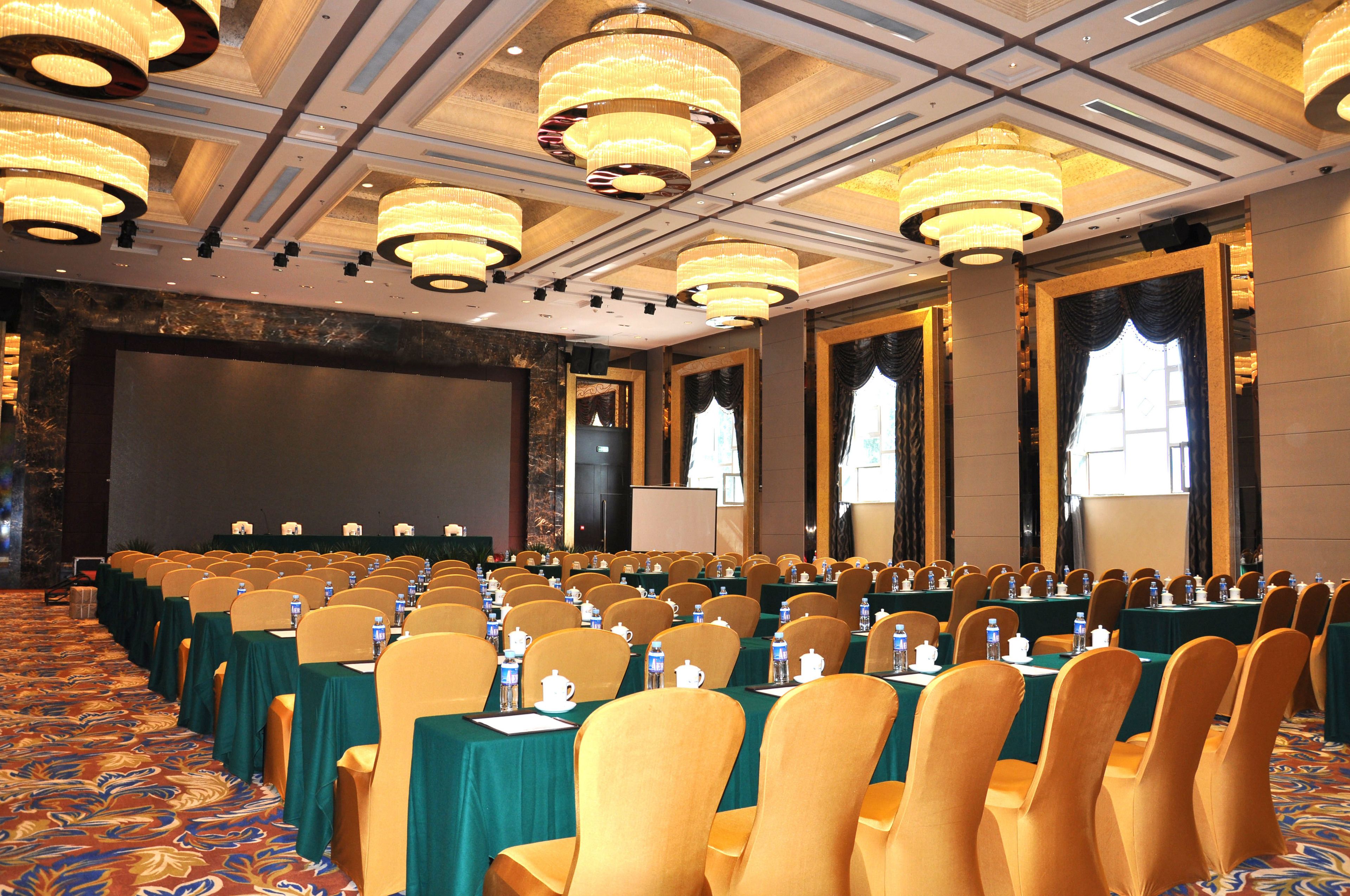 Un salón de conferencias con filas de mesas y sillas, grandes candelabros y una pantalla grande al frente.