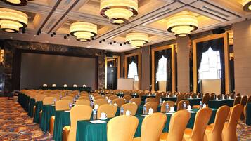 Un salón de conferencias con filas de mesas y sillas, grandes candelabros y una pantalla grande al frente.