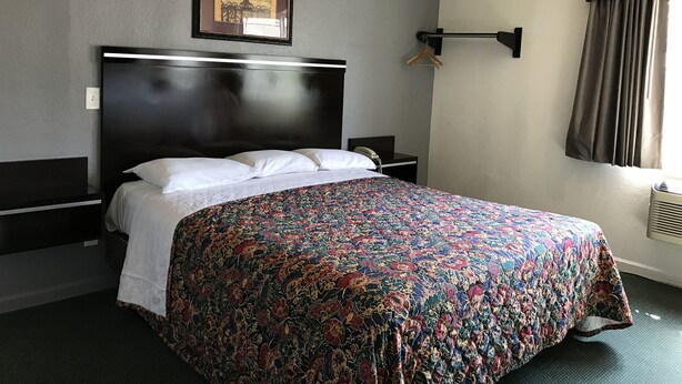 Ein Hotelzimmer mit einem großen Bett, einem Kopfteil, einem Nachttisch mit Lampe, einem gerahmten Bild an der Wand und einem Fenster mit Vorhängen.
