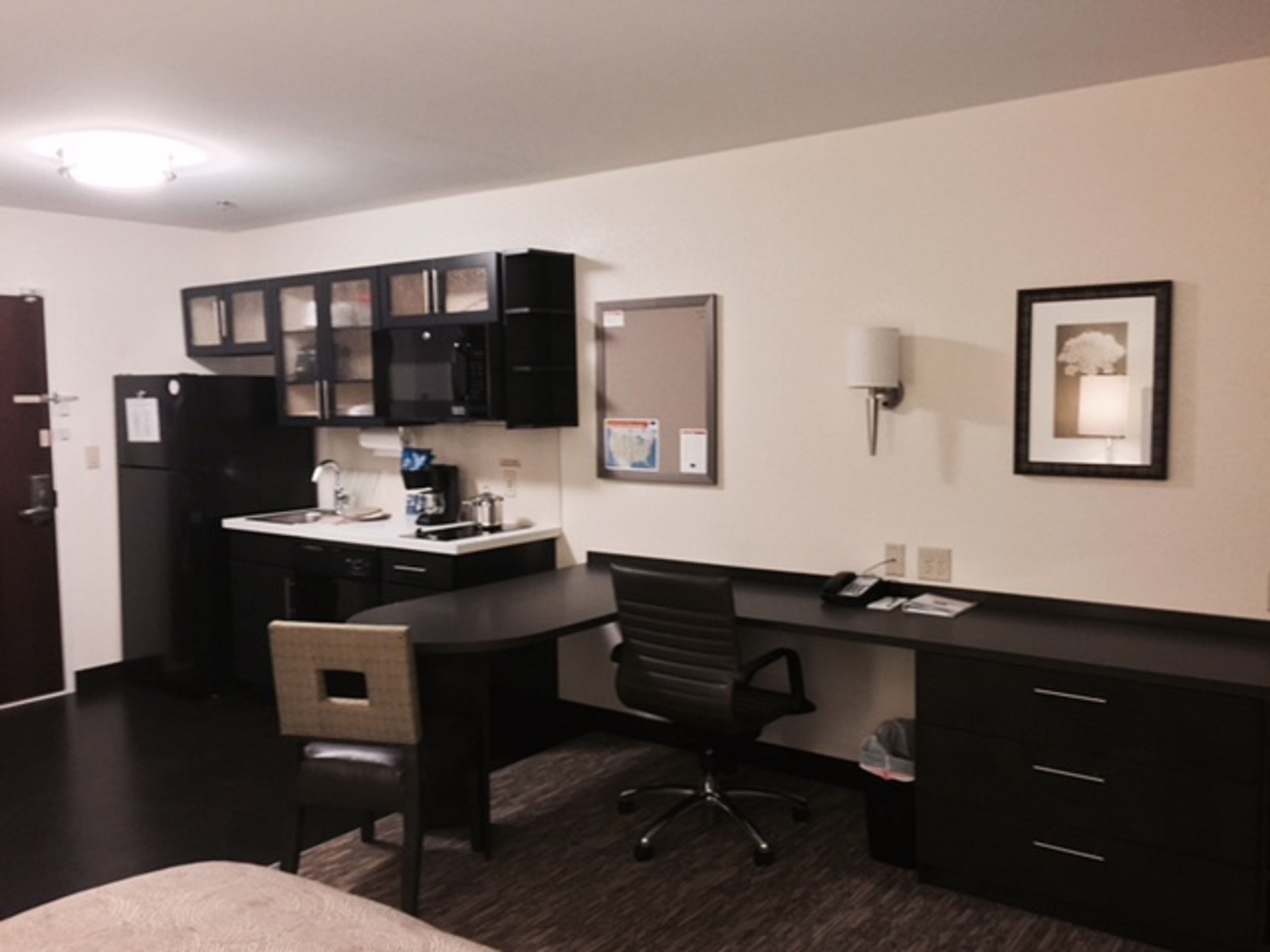 A hotel room with a kitchenette, a desk, a chair, a bed, and a framed picture on the wall.