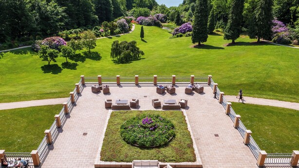 Una terraza ajardinada con zonas de estar y un jardín central, con vistas a un paisaje verde exuberante y una masa de agua.