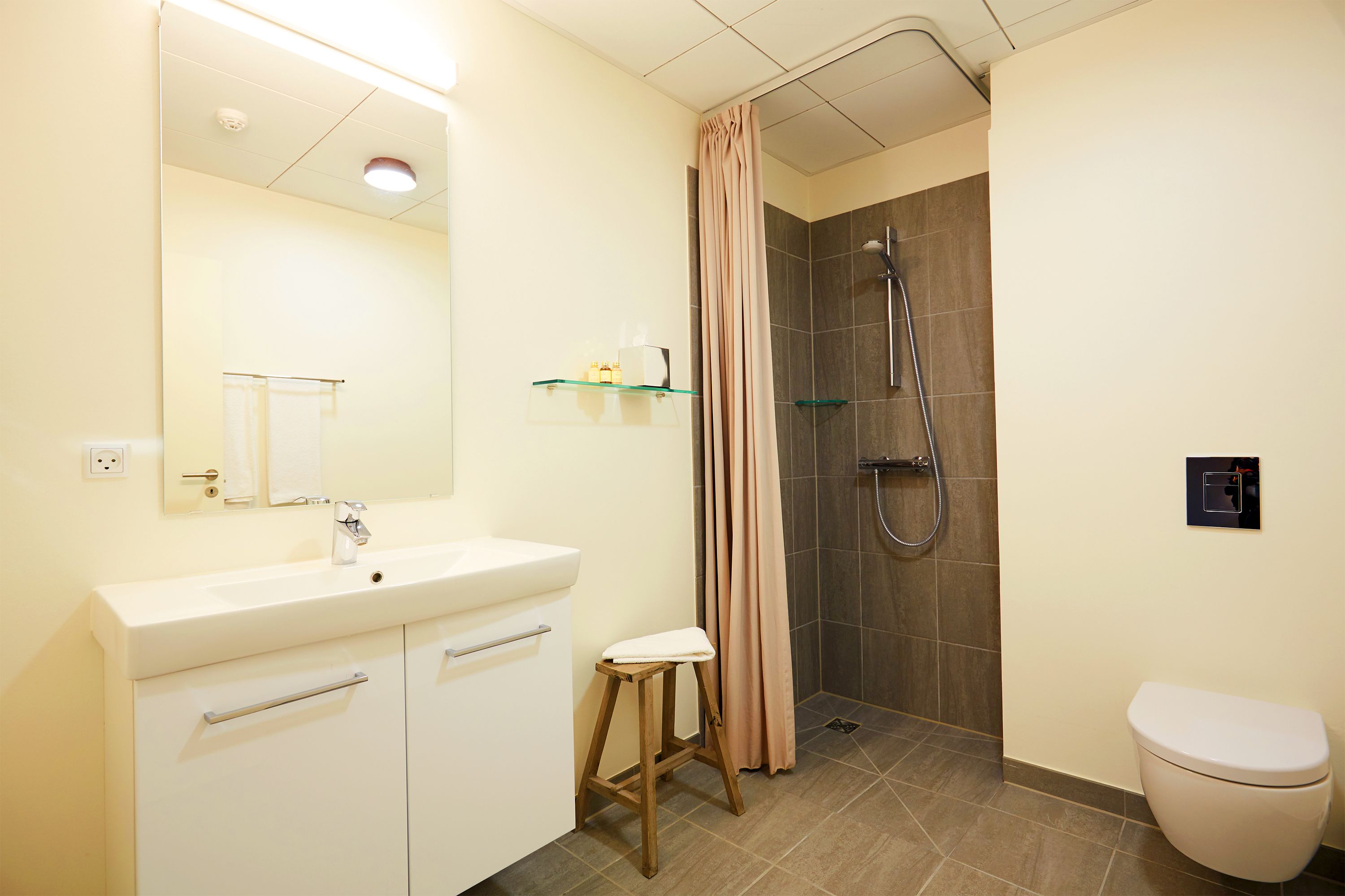 Une salle de bain avec un lavabo blanc, un miroir et des toilettes, une douche avec une porte en verre et un rideau beige.