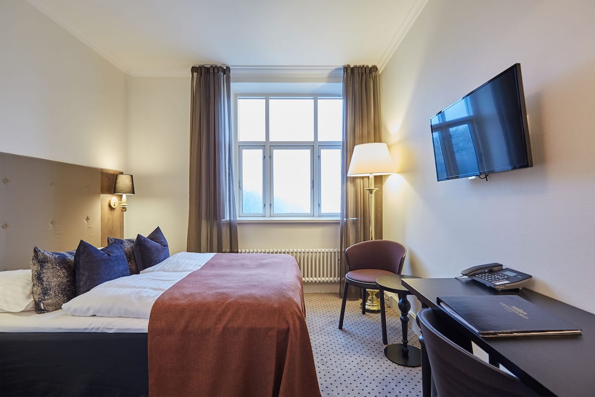 A hotel room with a bed, a desk with a chair, a TV, and a window with curtains.