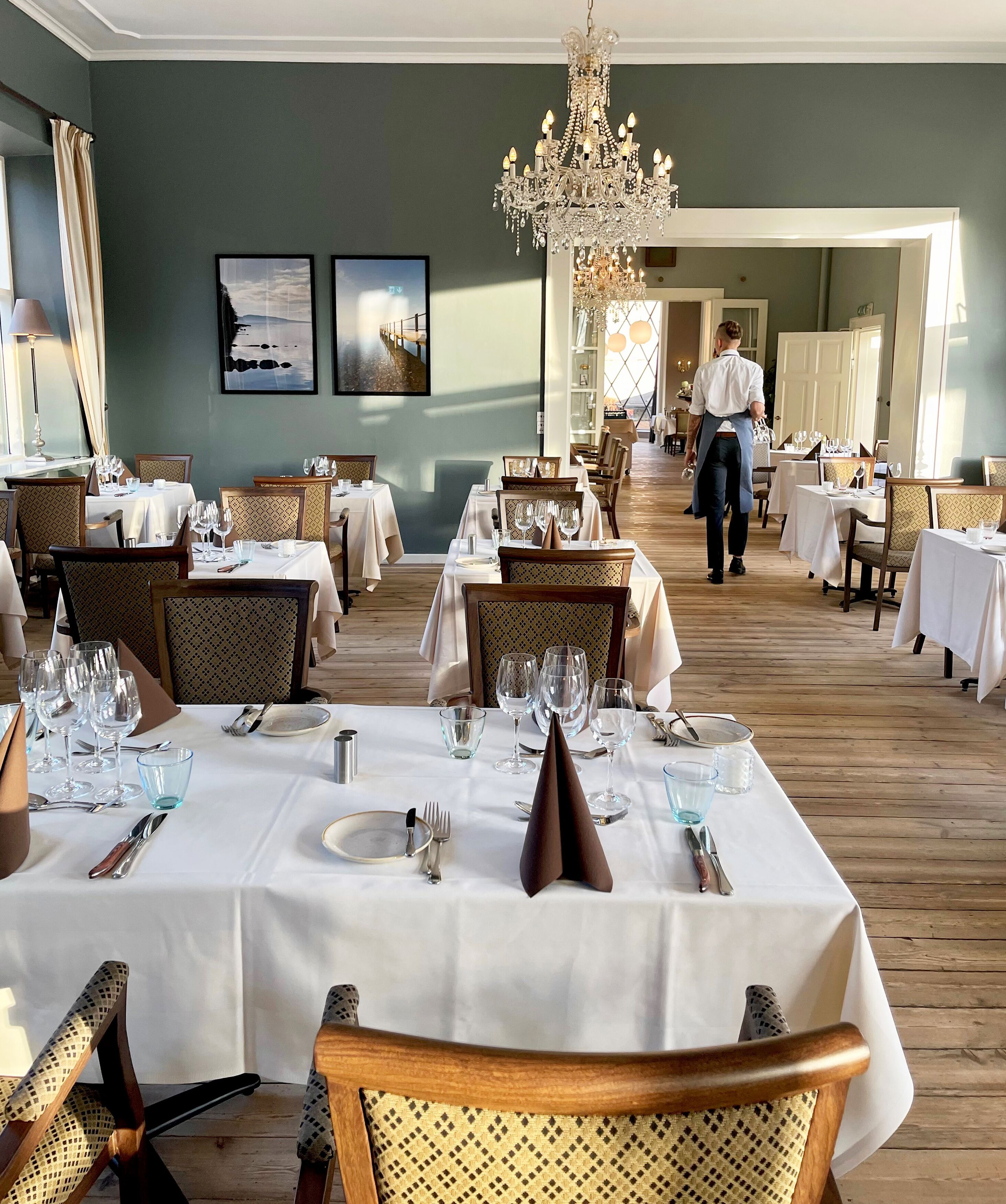 A restaurant with multiple tables set for dining, a waiter walking through, and a chandelier.