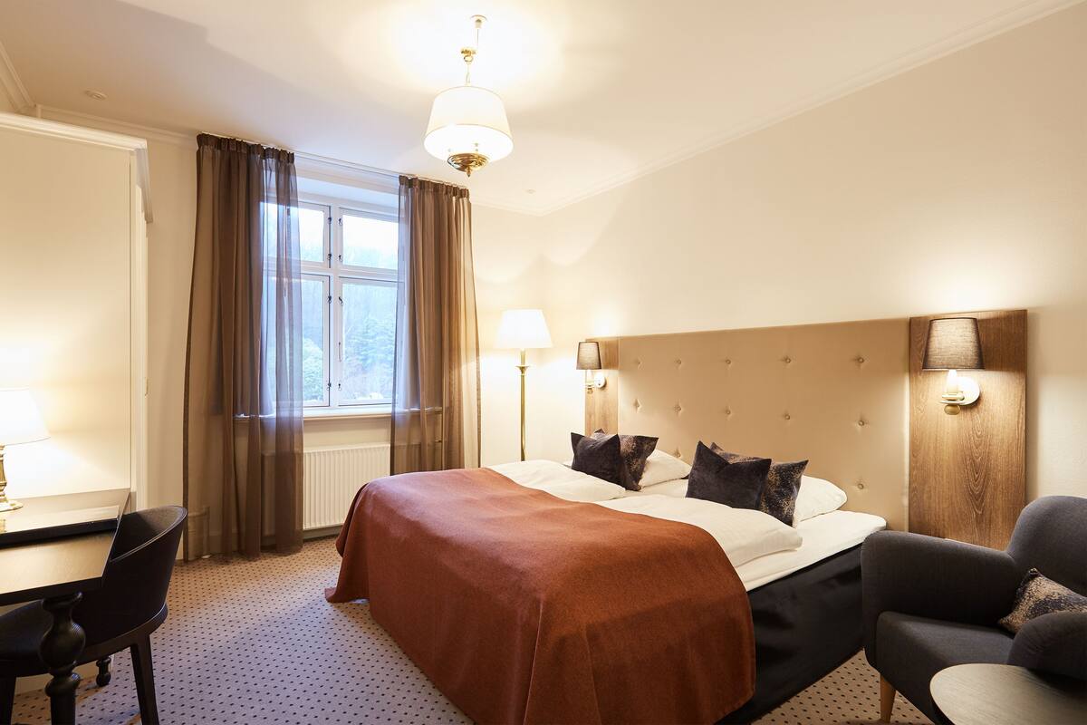 A hotel room with a large bed, a desk, and a chair. There is a window with curtains, a lamp, and a wall-mounted light fixture.