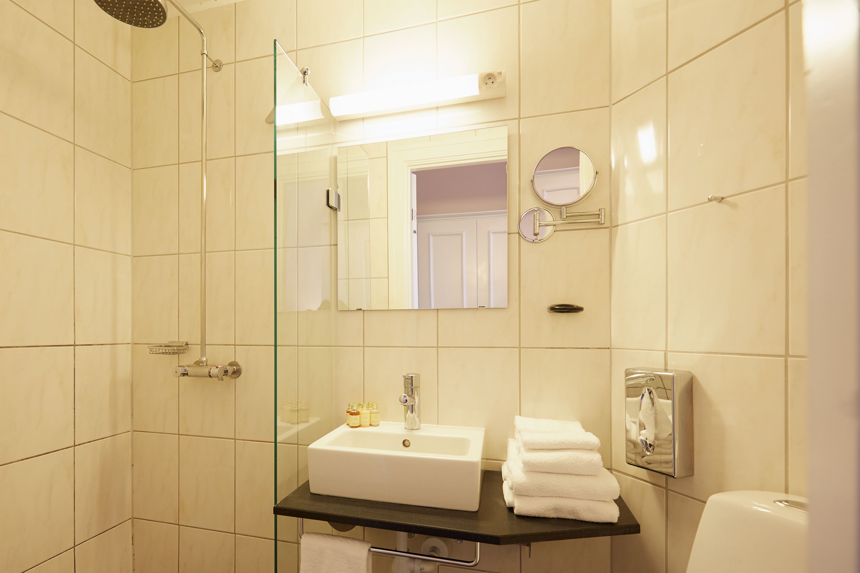 A bathroom with a glass shower enclosure, a white sink with a black countertop, a towel rack, and a toilet paper holder.