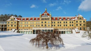 Un edificio grande e histórico con varios pisos y una torre central destacada, rodeado de nieve y árboles.