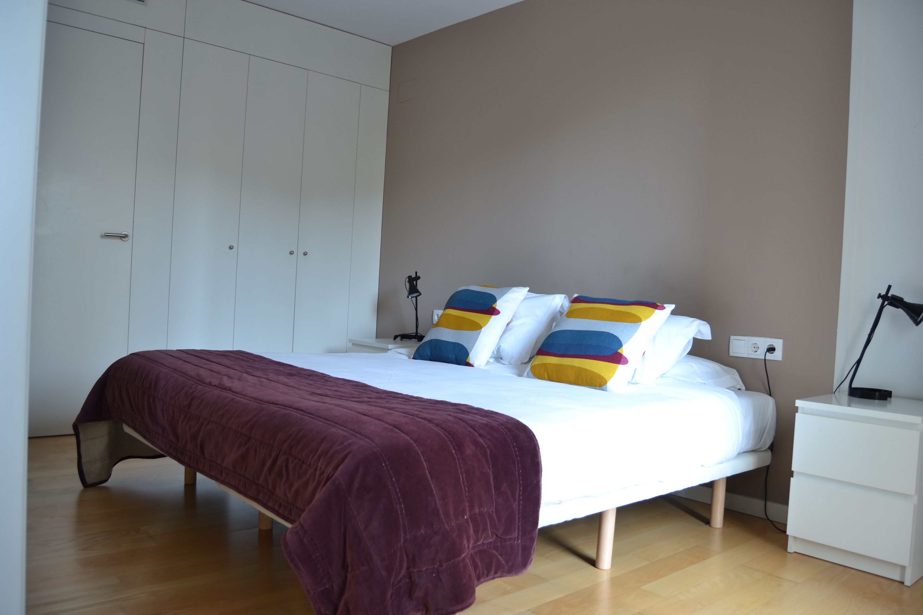 Un dormitorio con cama, mesitas de noche blancas, suelo de madera y armario.
