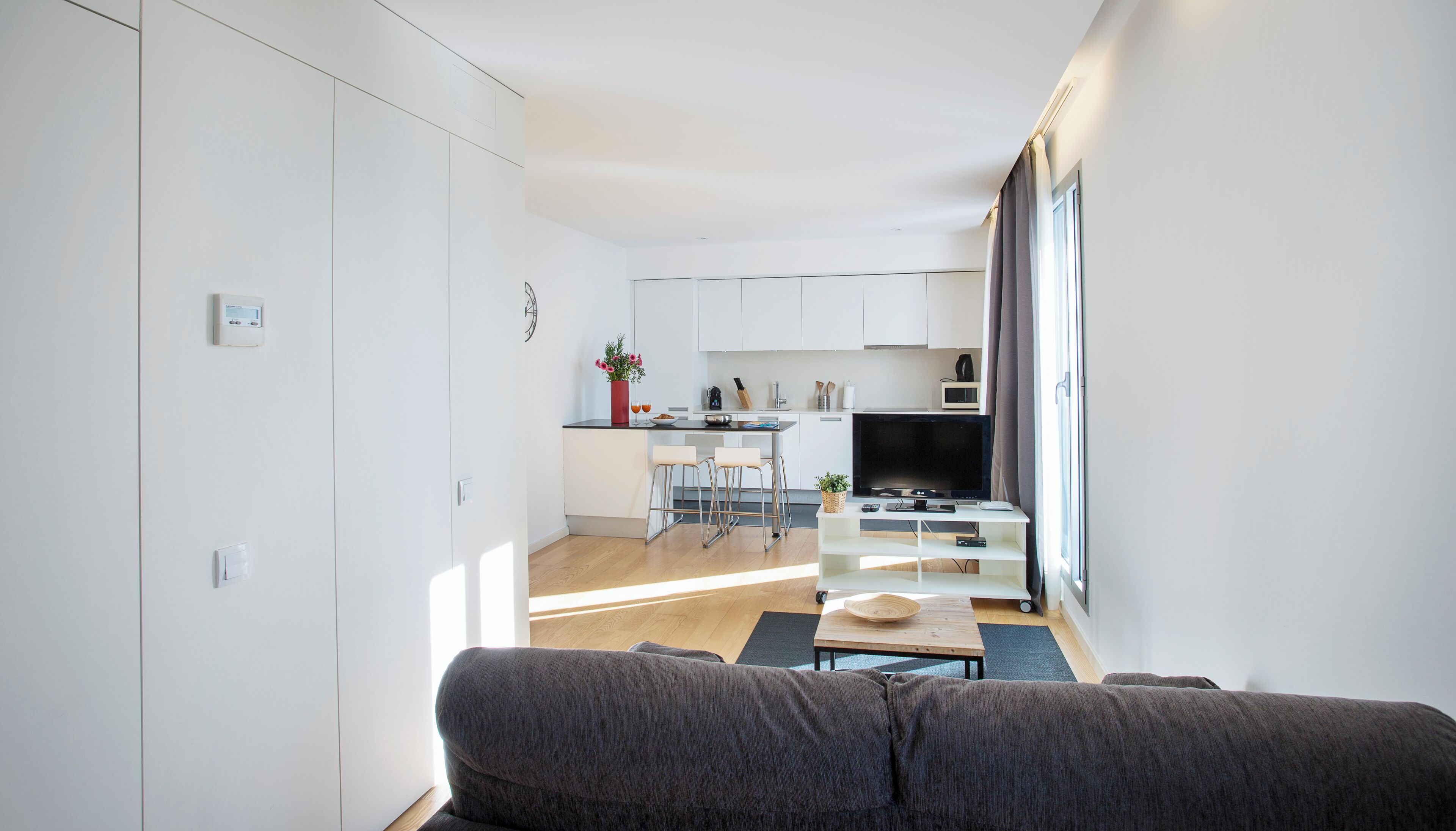 A modern living room with a grey sofa, a dining area with a small table and chairs, and a kitchen with white cabinets and a TV.