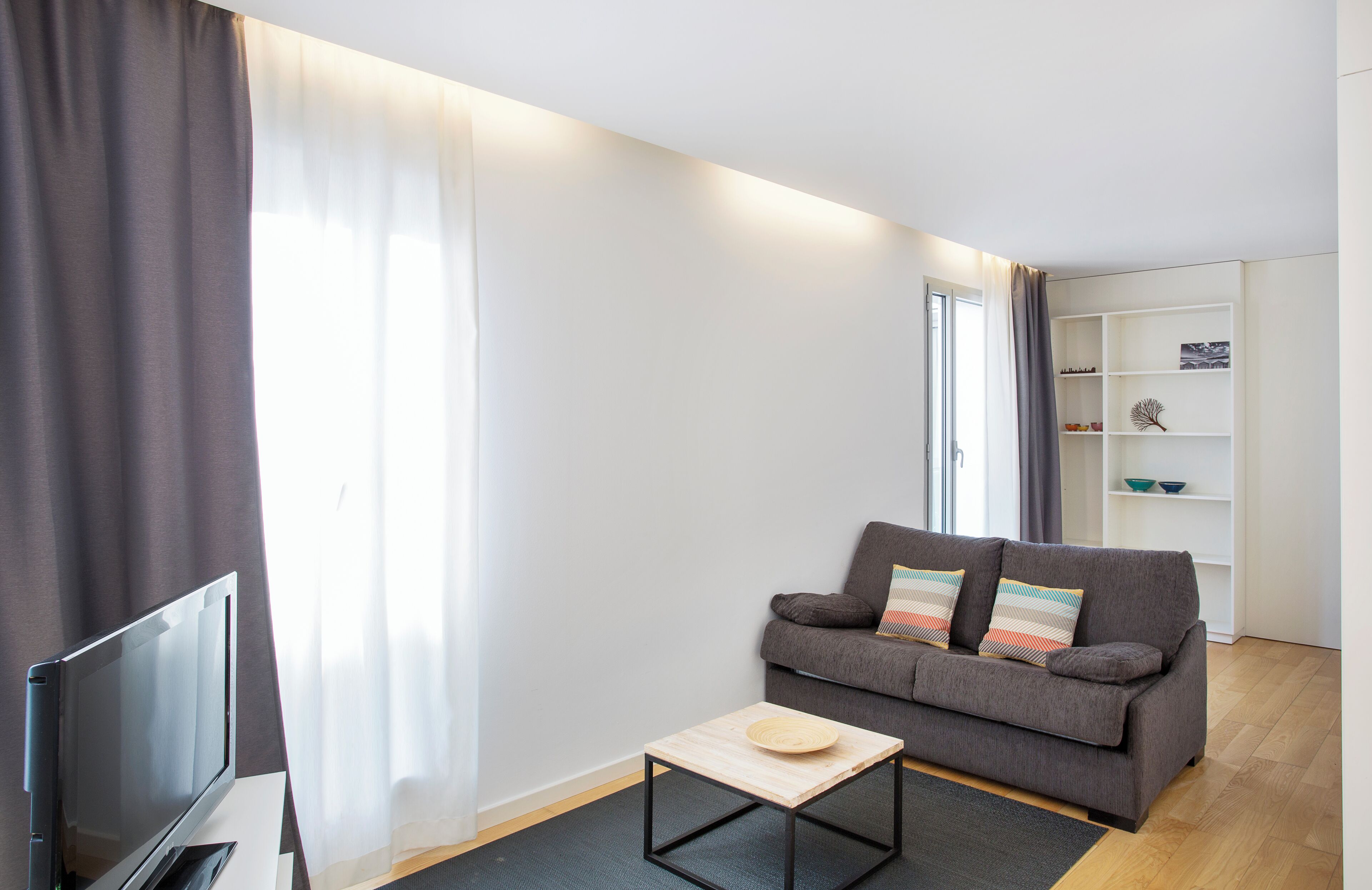 A modern living room with a grey sofa, a small wooden table, a TV, and a white shelving unit.