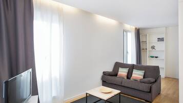A modern living room with a grey sofa, a small wooden table, a TV, and a white shelving unit.