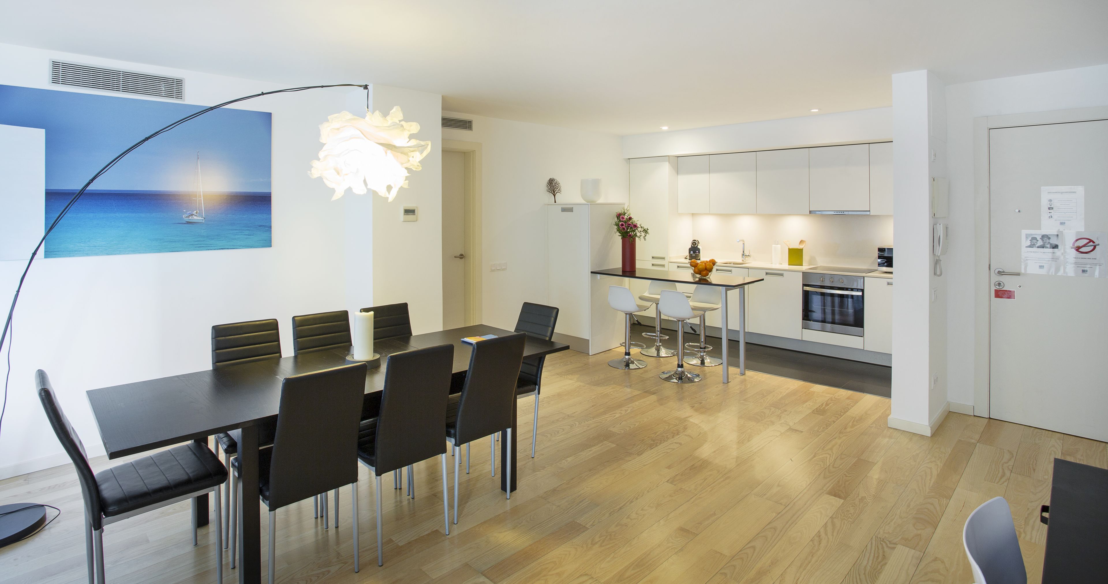 A modern kitchen with white cabinets, a dining area with a black table and chairs, and a painting of a sailboat on the wall.