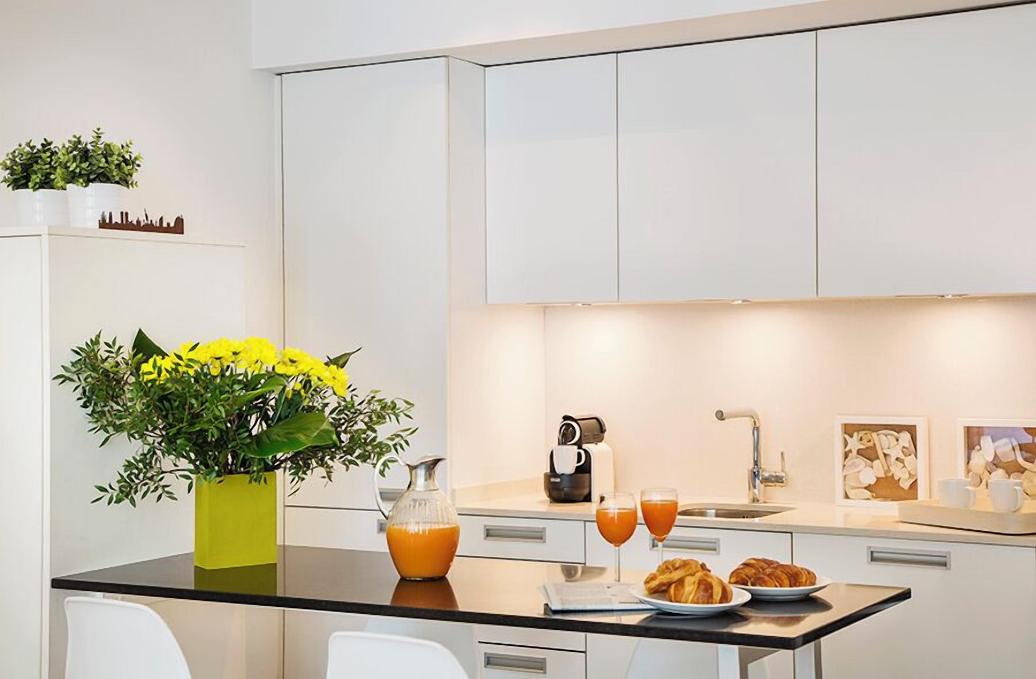 A modern kitchen with white cabinets, a black countertop, a coffee machine, two glasses of orange juice, and a plate of croissants.