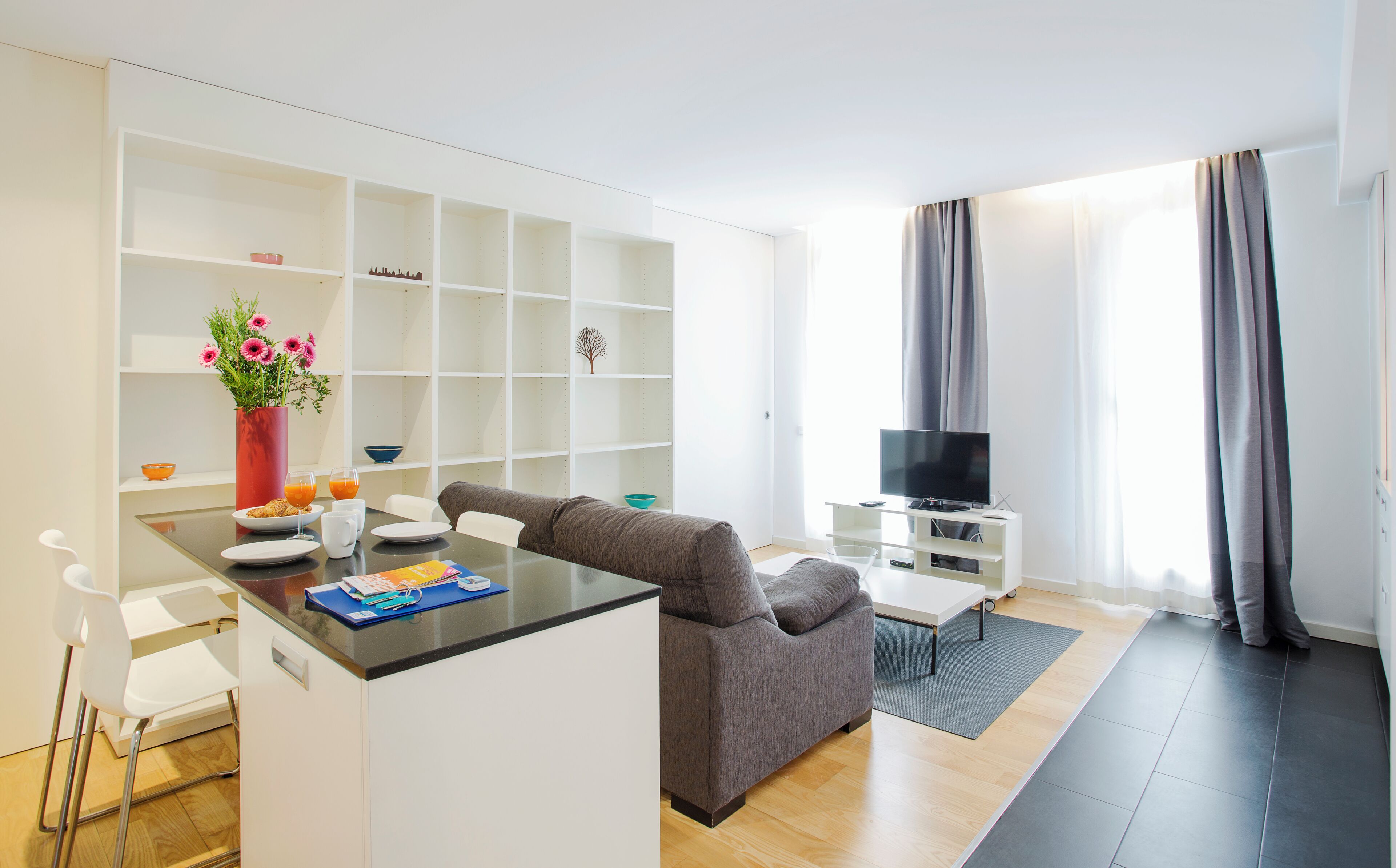 A modern living room with a grey sofa, a coffee table, and a TV on a stand. There is a white shelving unit with various items on the shelves.