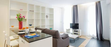 A modern living room with a grey sofa, a coffee table, and a TV on a stand. There is a white shelving unit with various items on the shelves.