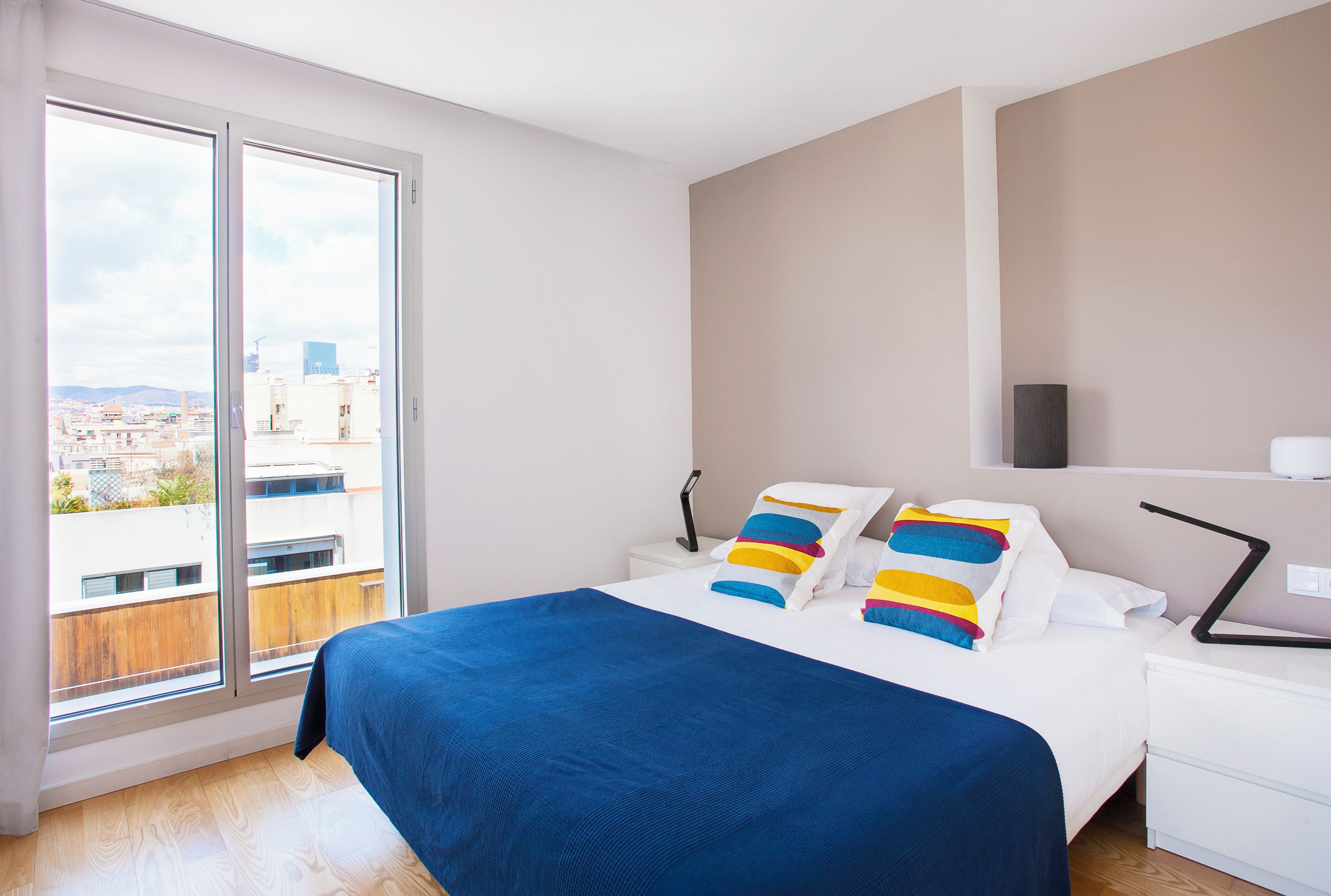 Un dormitorio moderno con una cama grande, una mesita de noche, un televisor y vistas de la ciudad a través de ventanales de piso a techo.