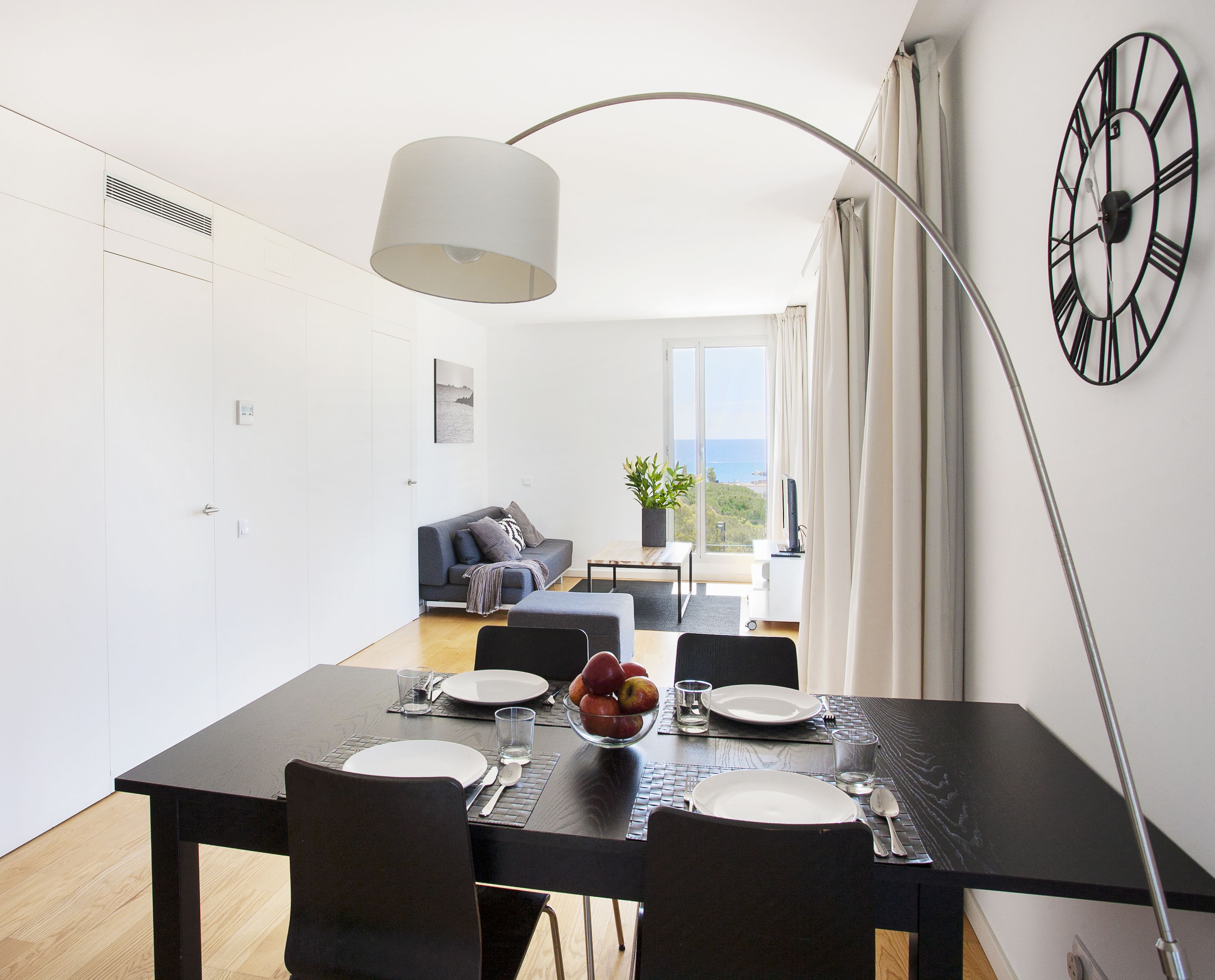A modern dining area with a black table set for four, a large clock on the wall, and a view of the sea through the window.