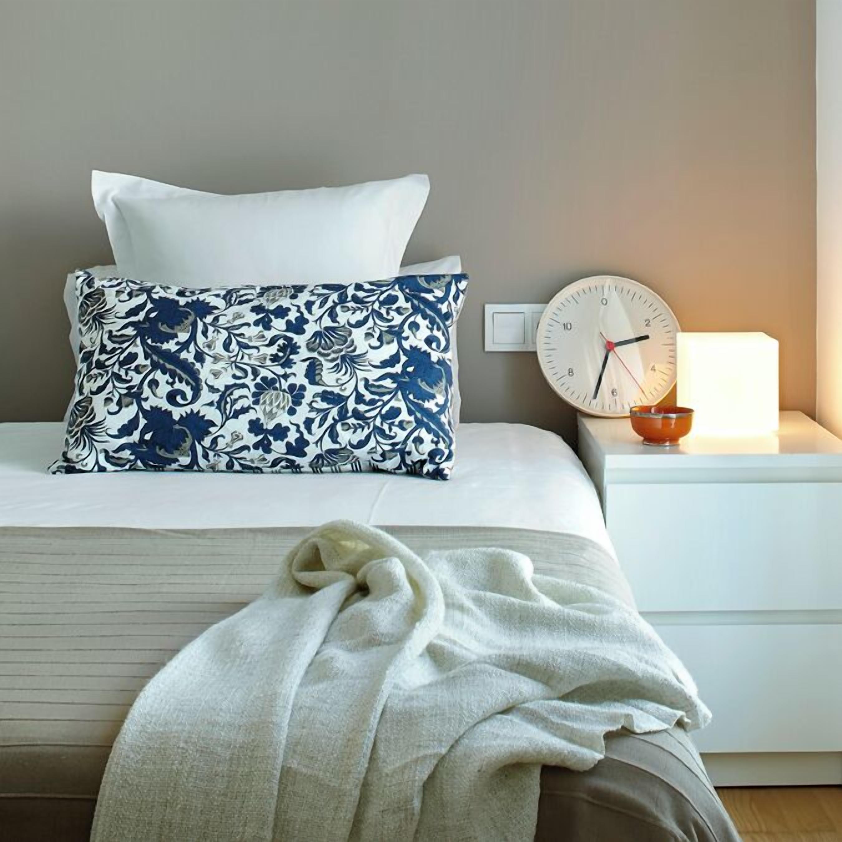 Una cama bien hecha con un almohadón blanco y azul estampada, una colcha blanca y un plaid blanco. Hay una mesita de noche con un reloj y un pequeño cuenco naranja.