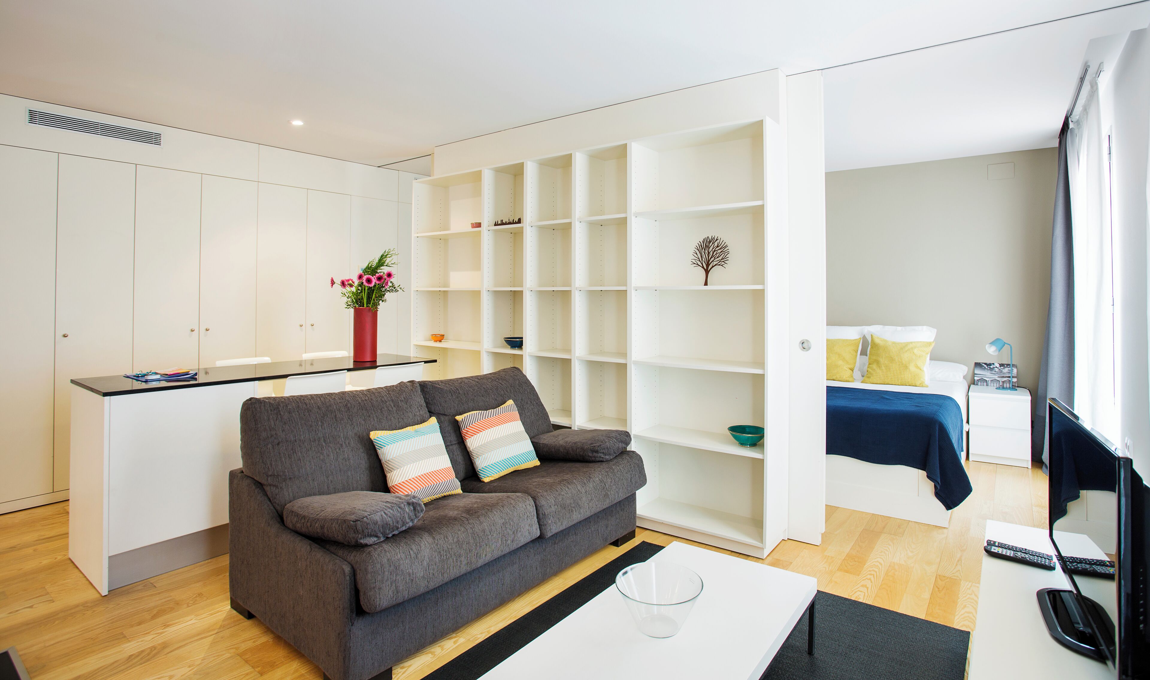 A modern living room with a grey sofa, a glass coffee table, and a white dining area with a red vase of flowers.
