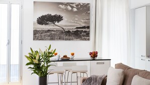 Ein modernes Wohnzimmer mit Couchtisch aus Holz, einem beigefarbenen Sofa, einer Essecke mit weißem Tisch und Stühlen sowie einem schwarz-weißen Landschaftsbild an der Wand.