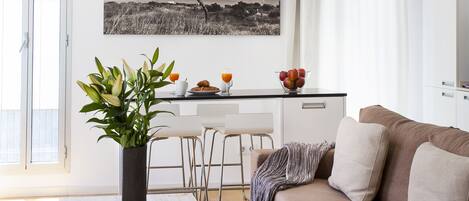 Una sala moderna con una mesa de centro de madera, un sofá color beige, una zona de comedor con una mesa y sillas blancas, y un cuadro de paisaje blanco y negro en la pared.