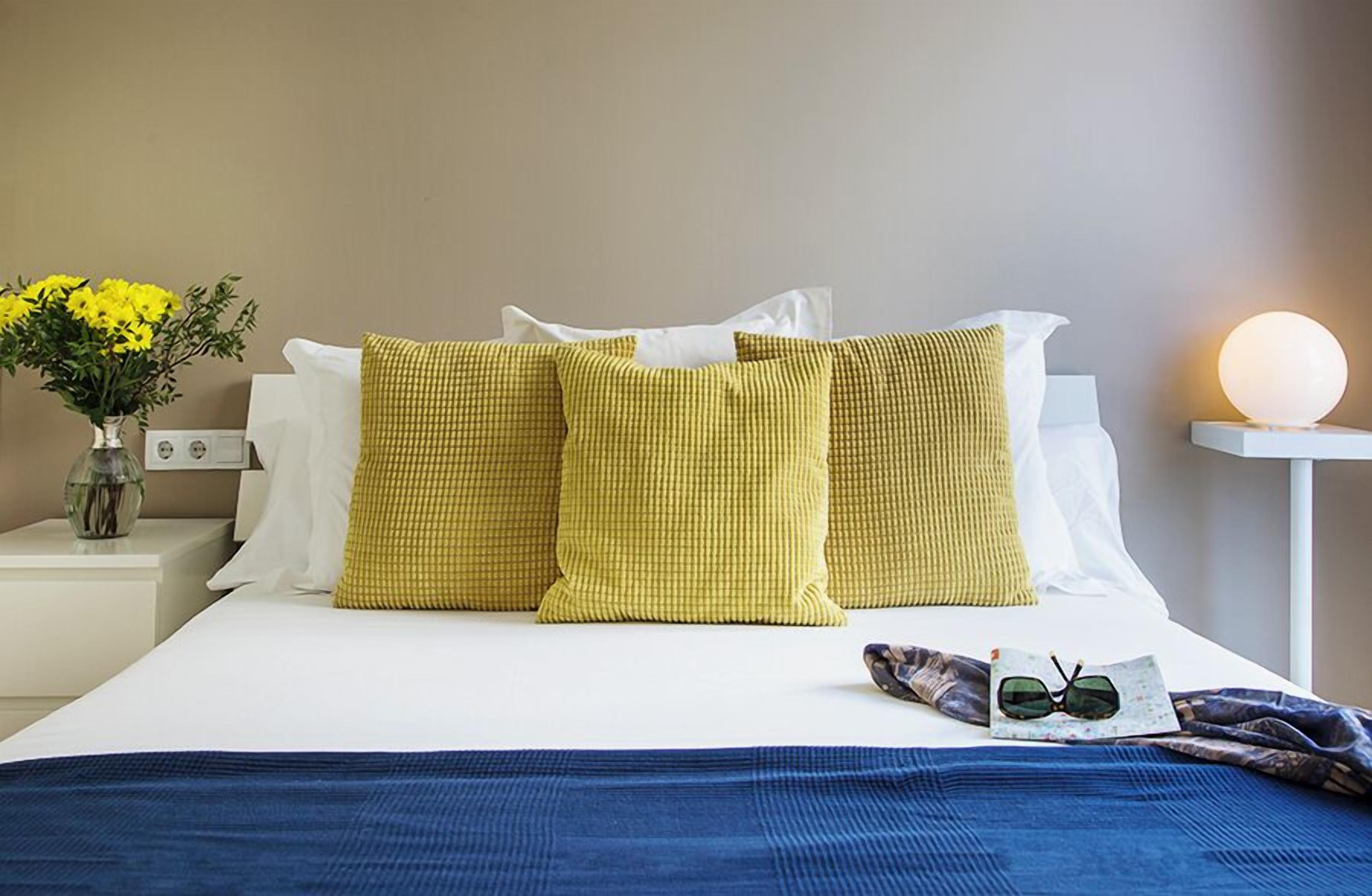 Ein ordentlich bezogenes Bett mit weißen und gelben Kissen, einer blauen Decke und einer Sonnenbrille darauf.