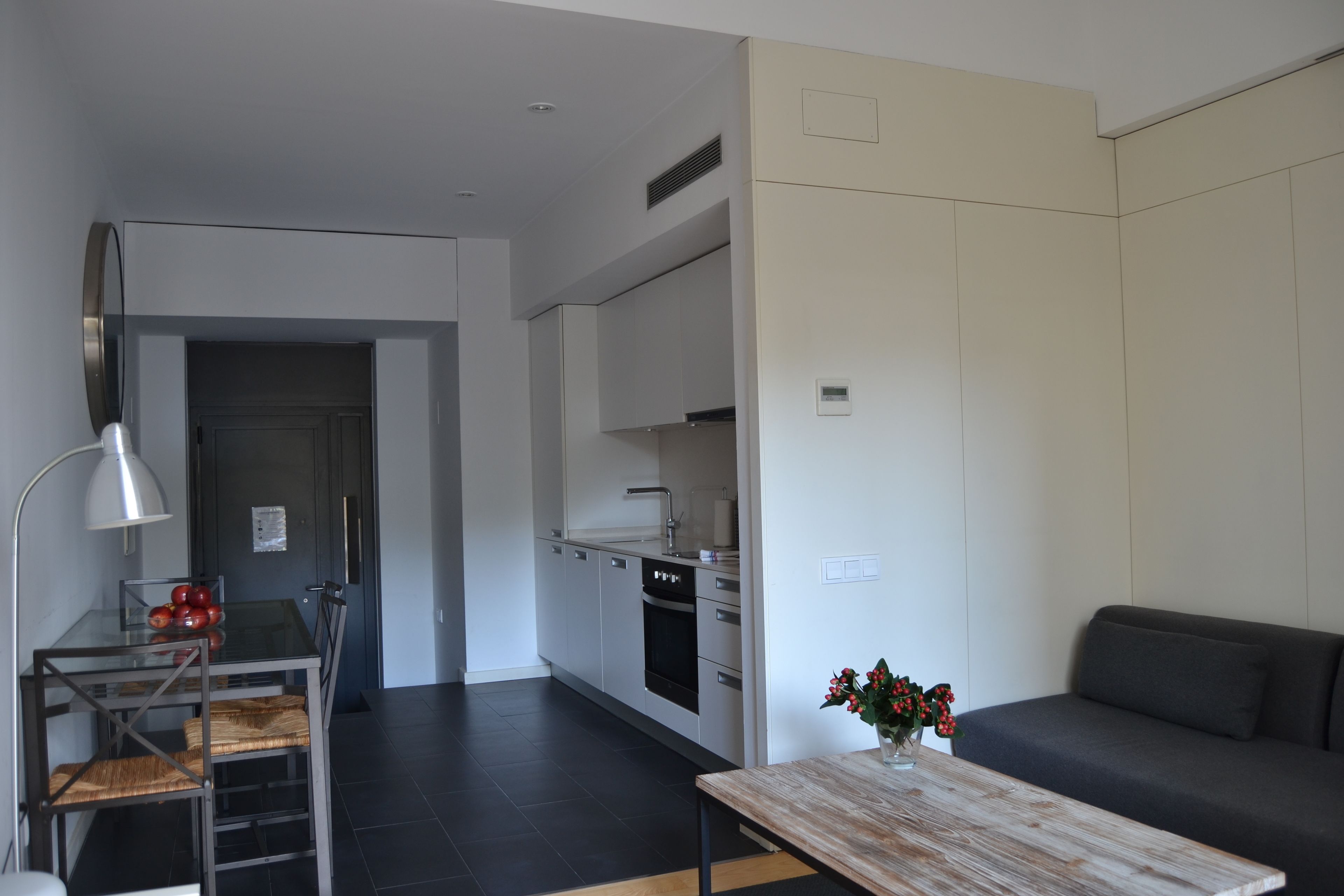 A modern living room with a glass dining table, a sofa, and a kitchen area with white cabinets and a black oven.