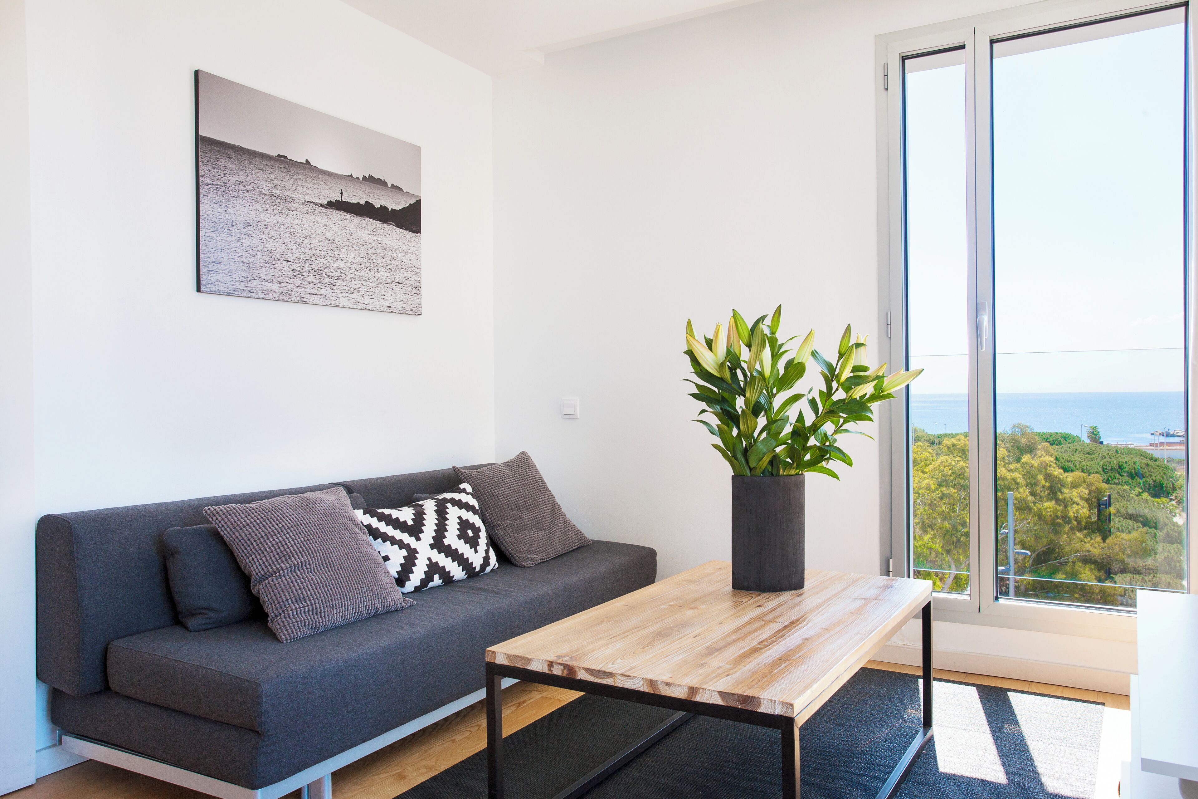 A modern living room with a grey sofa, a wooden coffee table, a potted plant, and a large window offering a view of the sea and greenery.