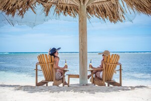 Dos sillas de madera bajo un toldo de paja en una playa con vista despejada al océano.