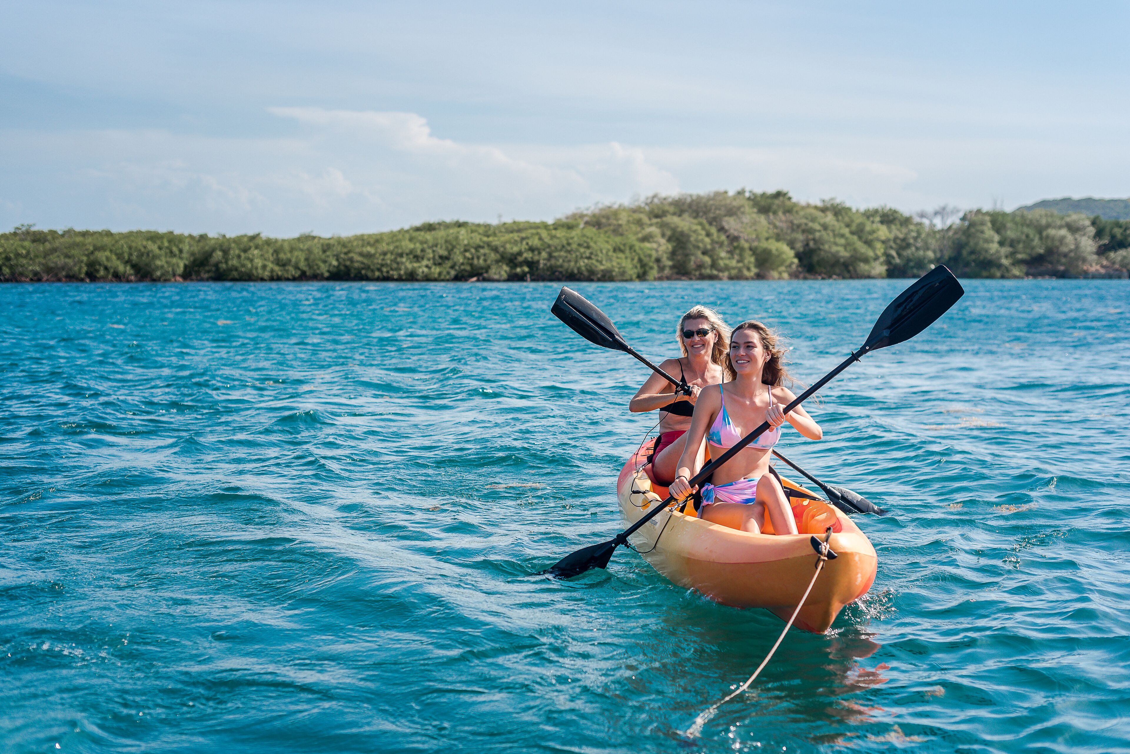 Dos personas en un bote inflable pequeño sobre un cuerpo de agua, con una orilla boscosa al fondo.