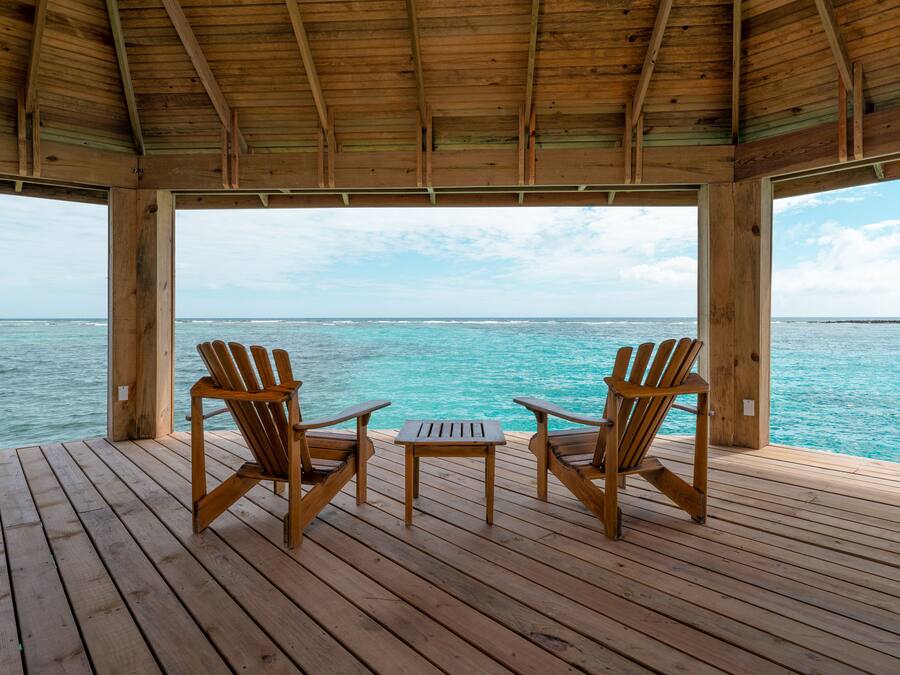 Un pontile in legno con due sedie e un tavolino, con vista su acqua turchese sotto un tetto in legno.