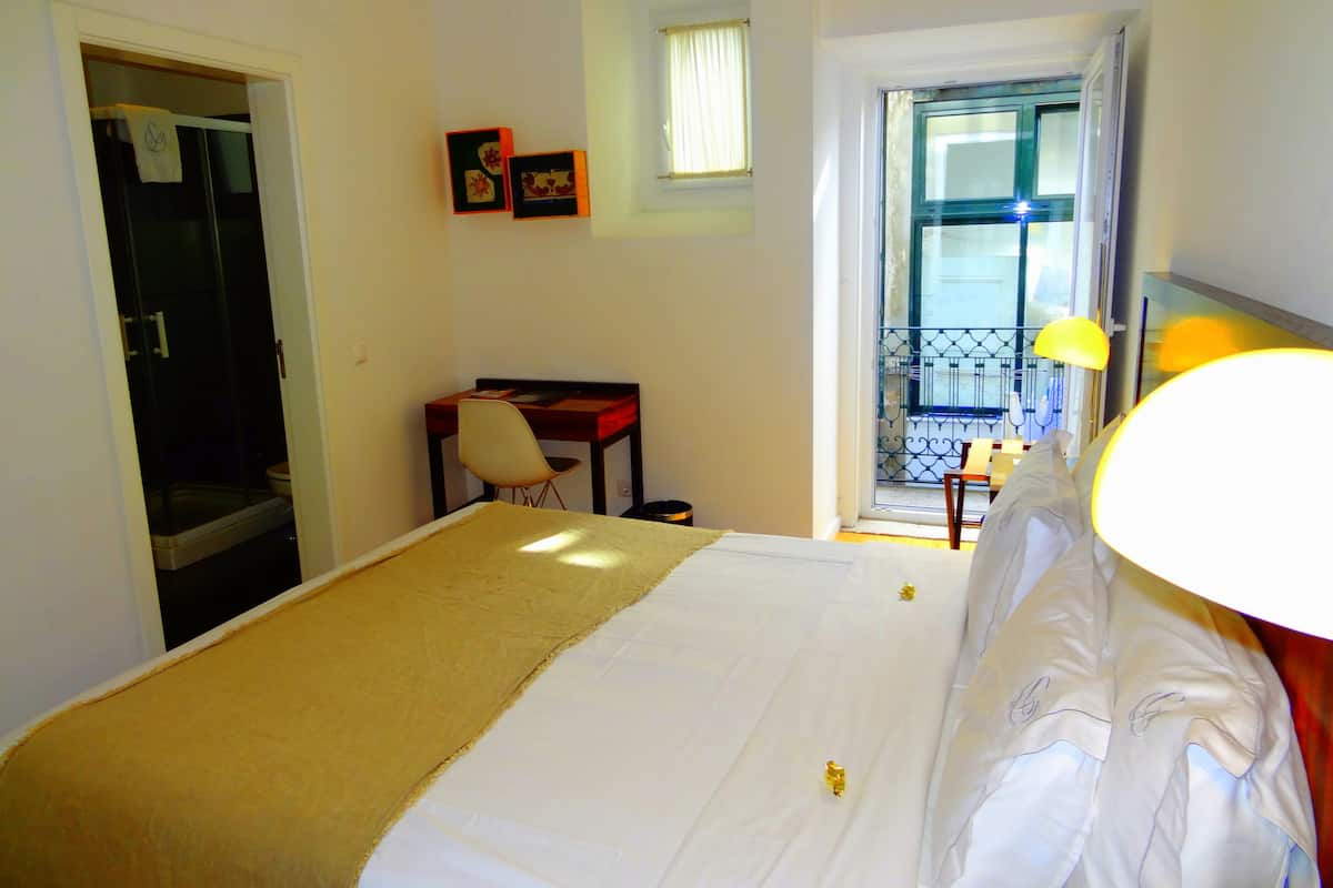 A hotel room with a bed, a desk, a chair, a window with a view, and a balcony with a railing.