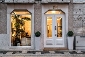 Entrada de hotel con puertas dobles, placa con número y plantas en macetas.