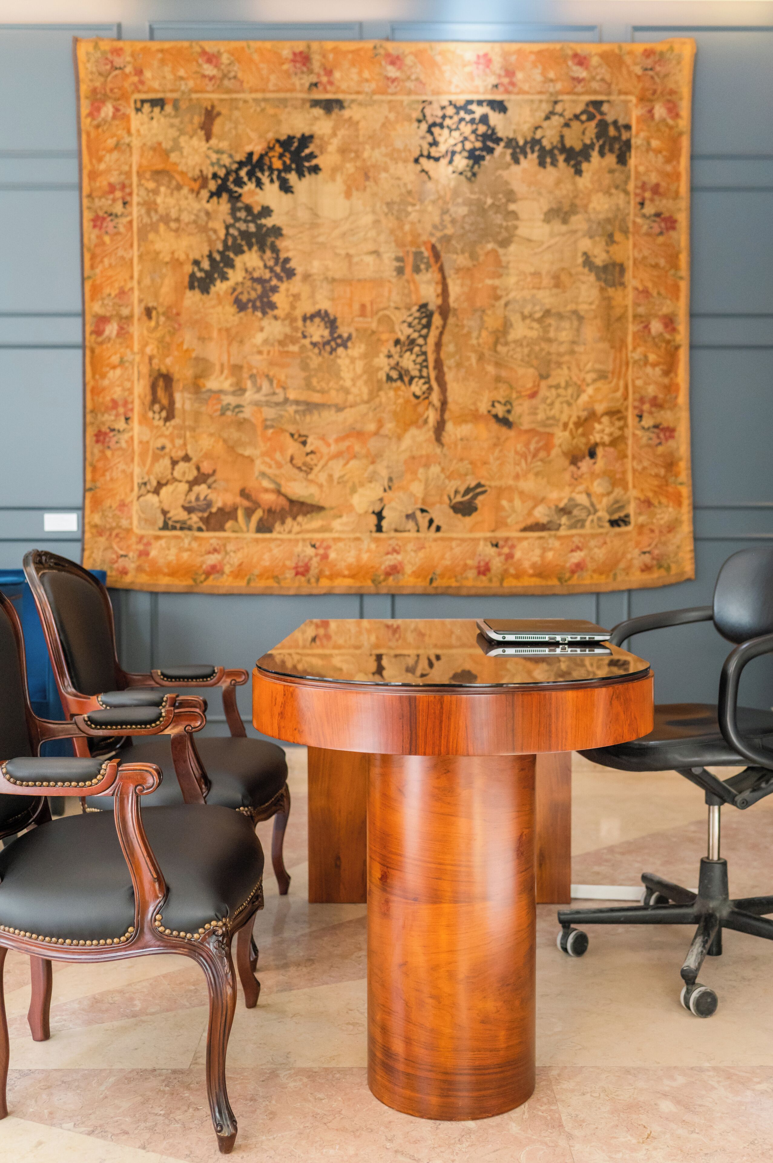 Una sala de conferencias con una mesa redonda de madera, dos sillones de cuero y un tapiz floral grande en la pared.