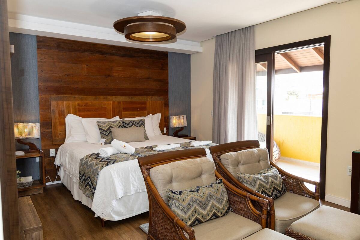 Habitación de hotel con una cama grande, dos mesitas de noche, un cabecero de madera, sillas de mimbre y un balcón con vistas.