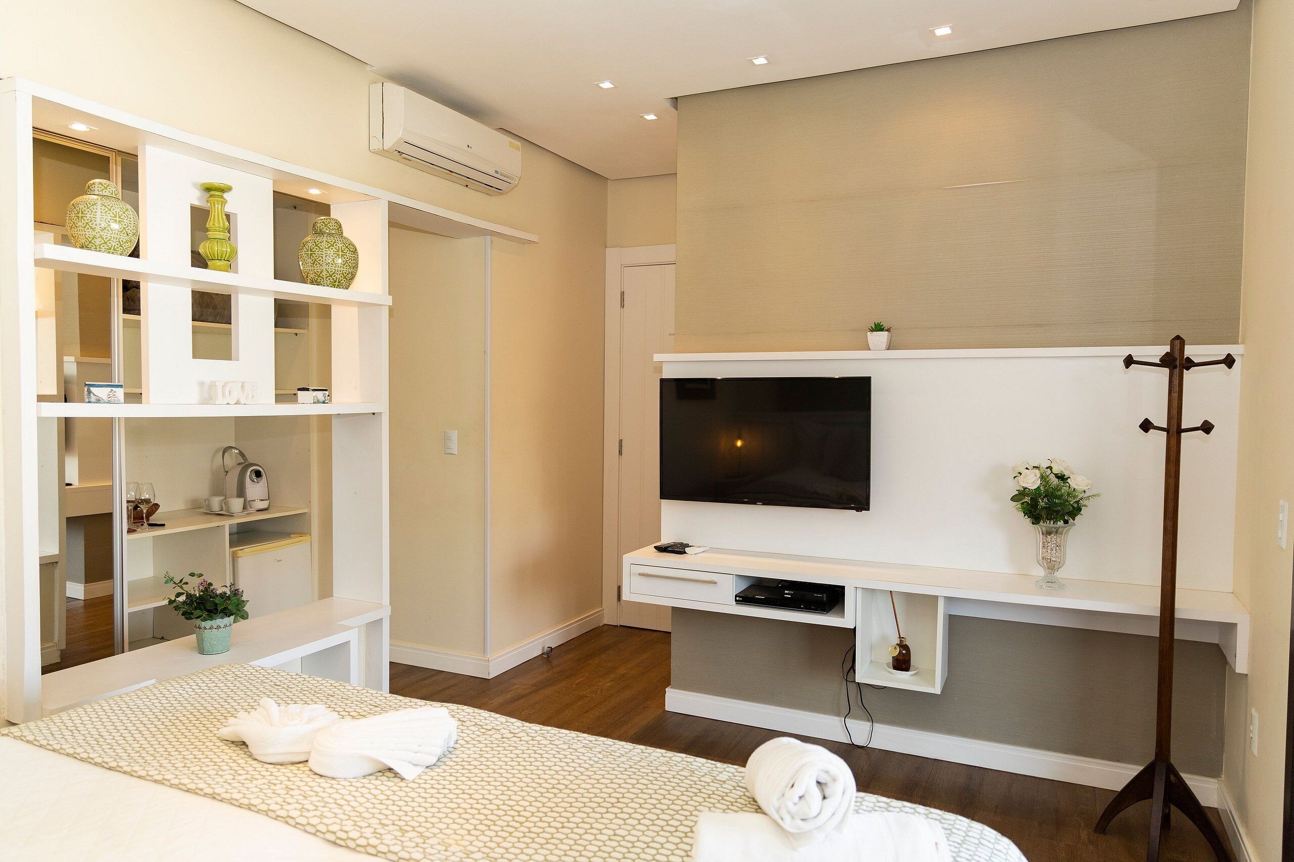 Una camera d'albergo con un letto, una TV a parete, uno scaffale con oggetti decorativi, un appendiabiti e una piccola pianta.