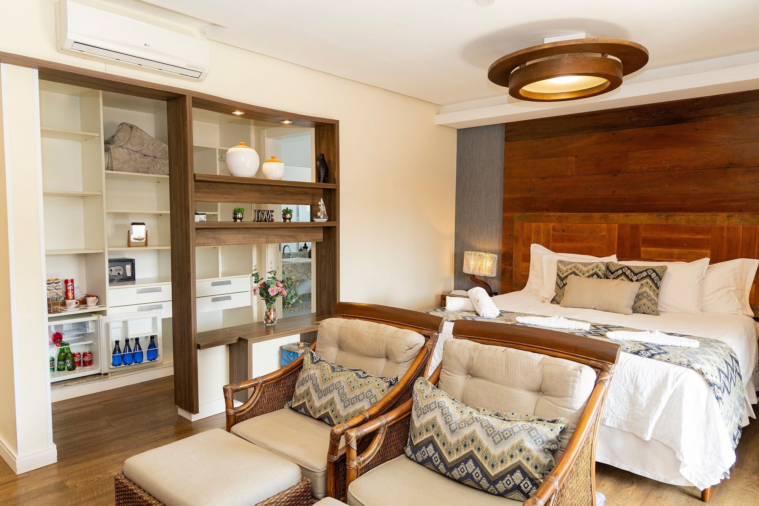 A hotel room with a bed, two wicker chairs, a wooden headboard, a built-in shelf unit, and a ceiling light fixture.