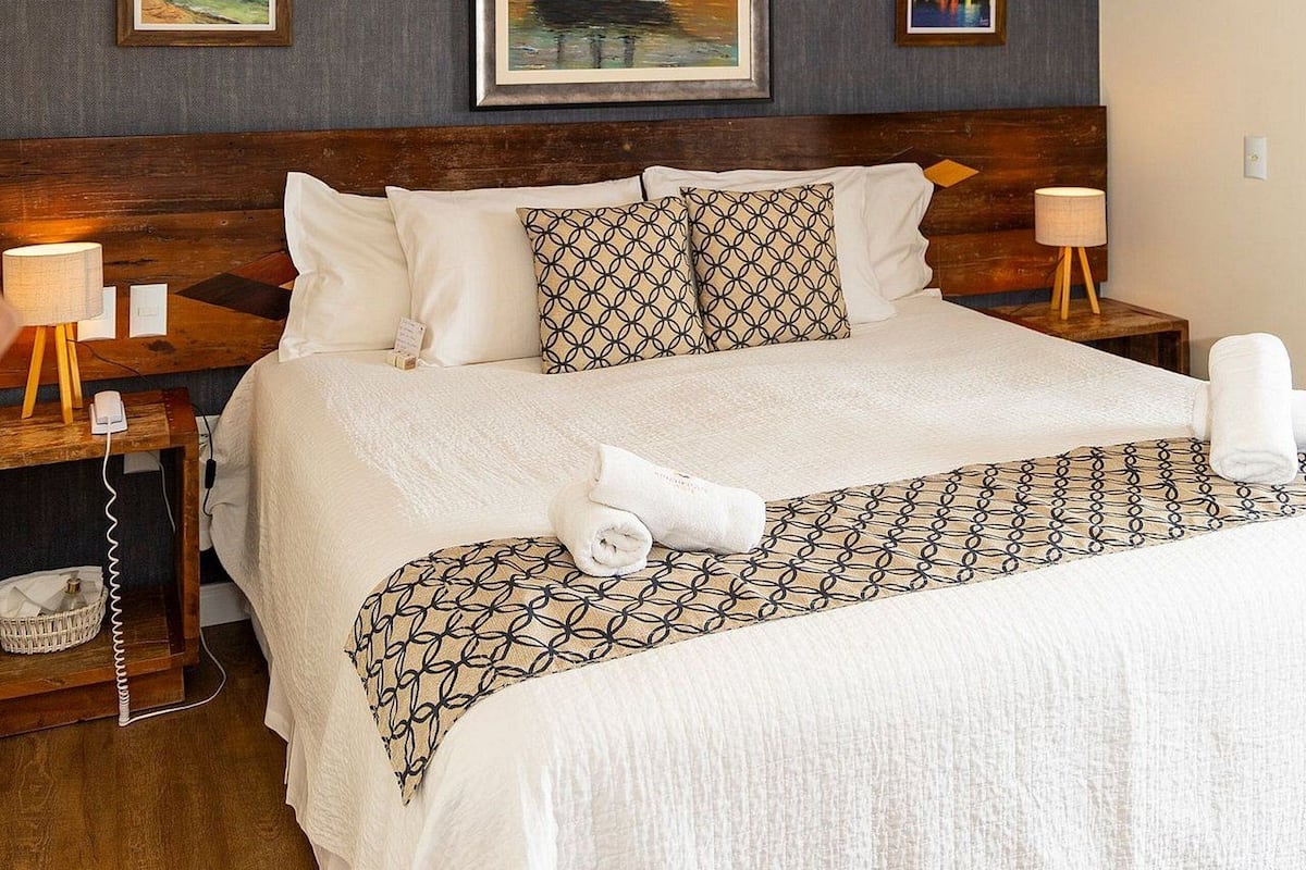 Una habitación de hotel con una cama grande, cabecera de madera, un cuadro de un velero y una lámpara de techo moderna.