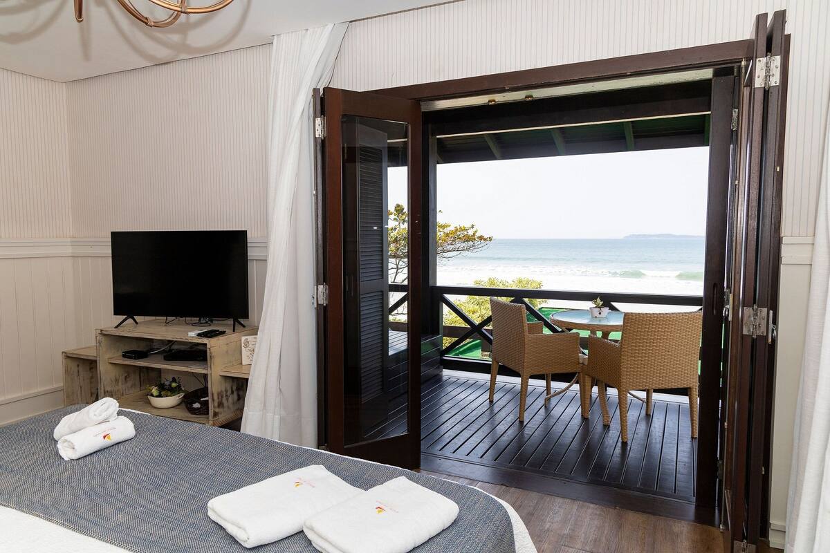Habitación de hotel con cama, televisor, balcón con mesa y sillas, y vista al océano.