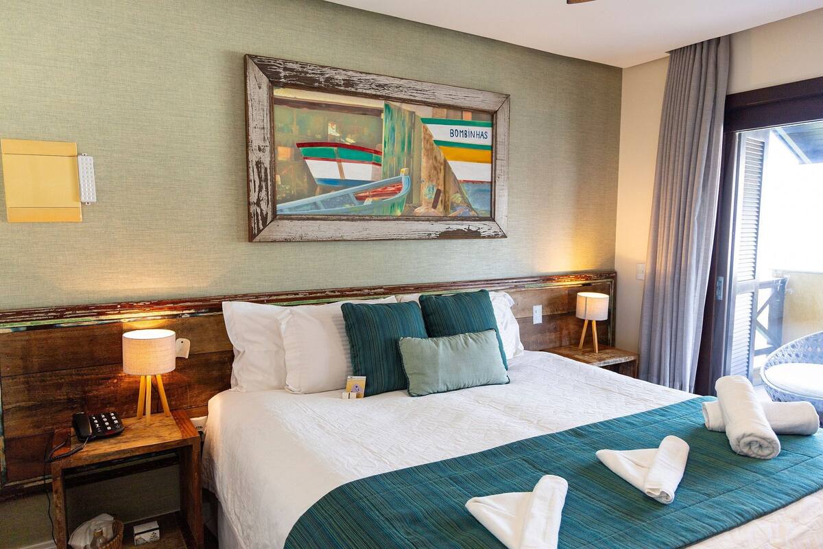 Habitación de hotel con cama, mesita de noche con lámpara, un cuadro en la pared y vista al exterior.