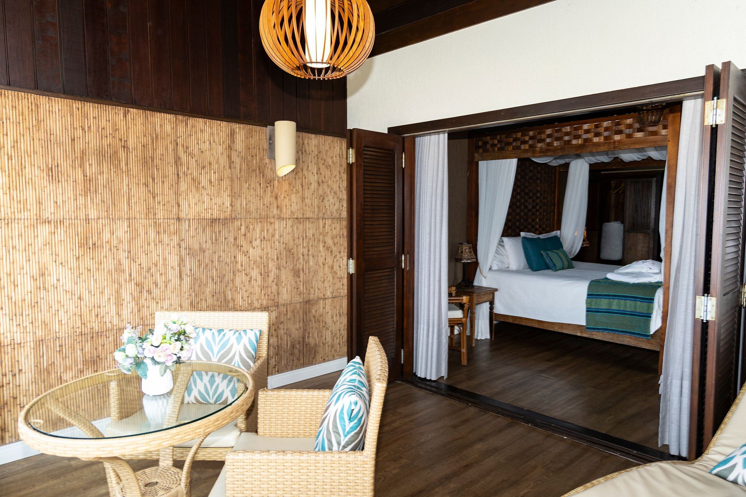 Una habitación con una cama, una mesita de vidrio, muebles de mimbre y techo de madera.