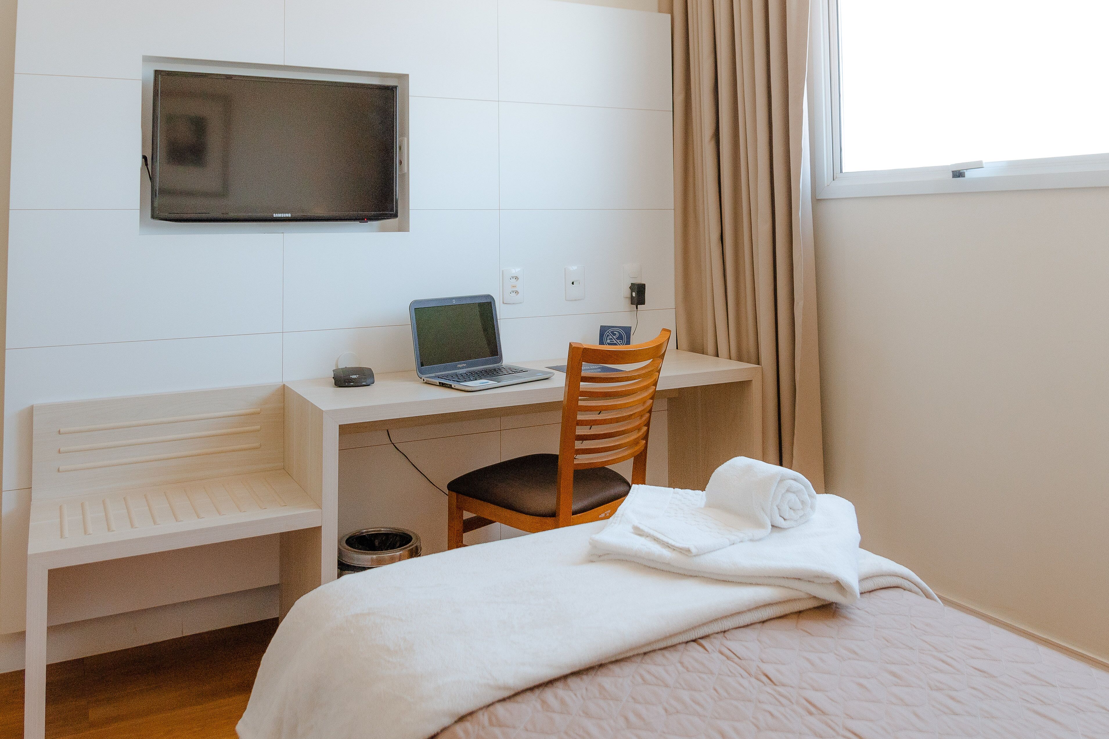 Habitación de hotel con cama, escritorio con computadora portátil, una silla, televisor montado en la pared y ventana con cortinas.