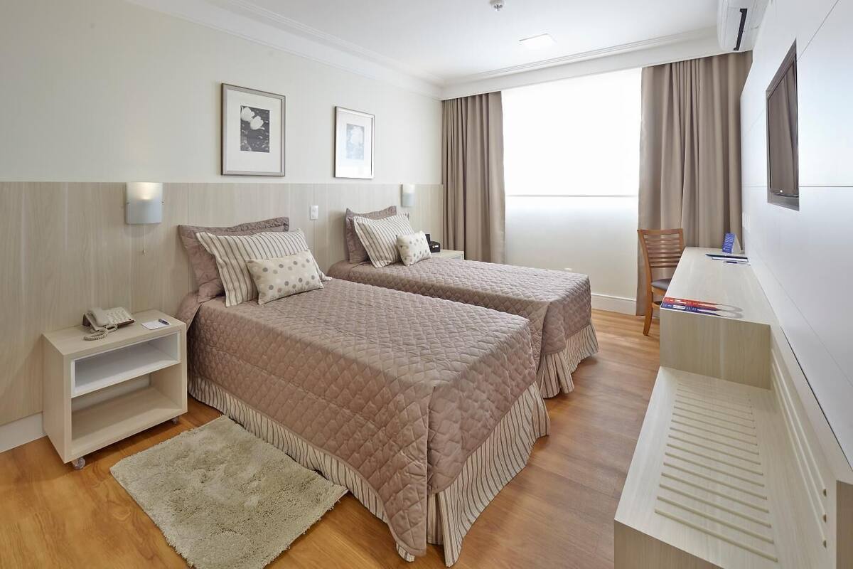 A hotel room with two beds, a desk, a chair, a TV, and a window with curtains.