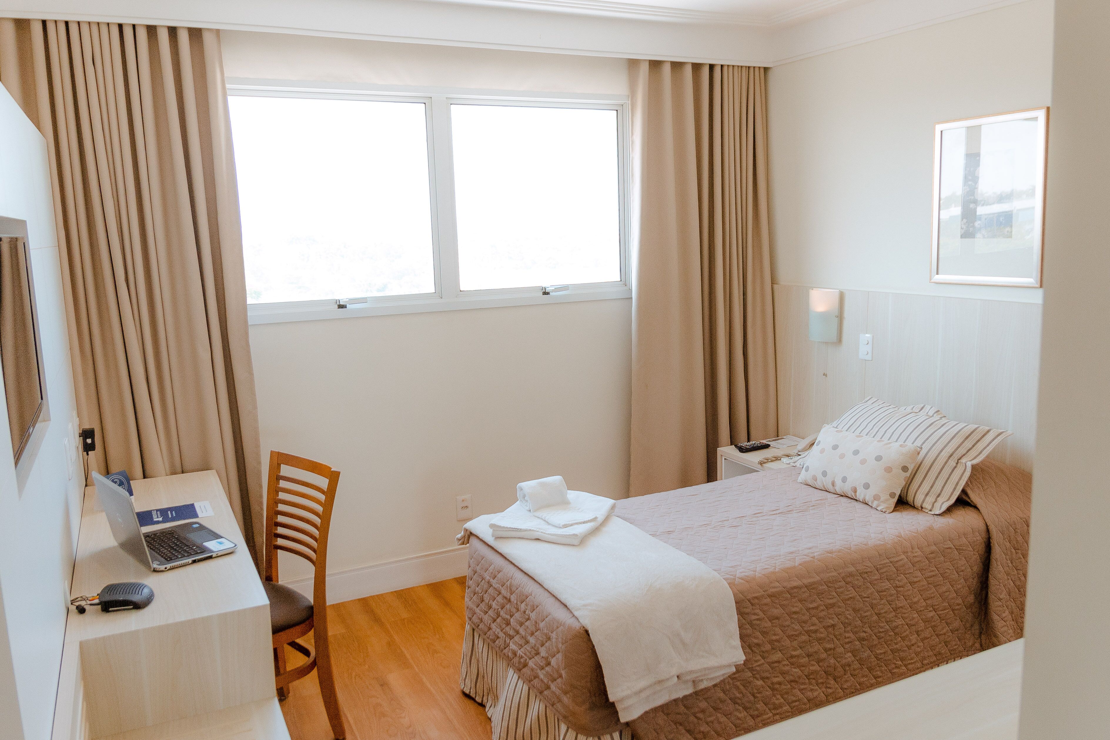 A hotel room with a bed, a desk with a laptop, a chair, and a window with curtains.