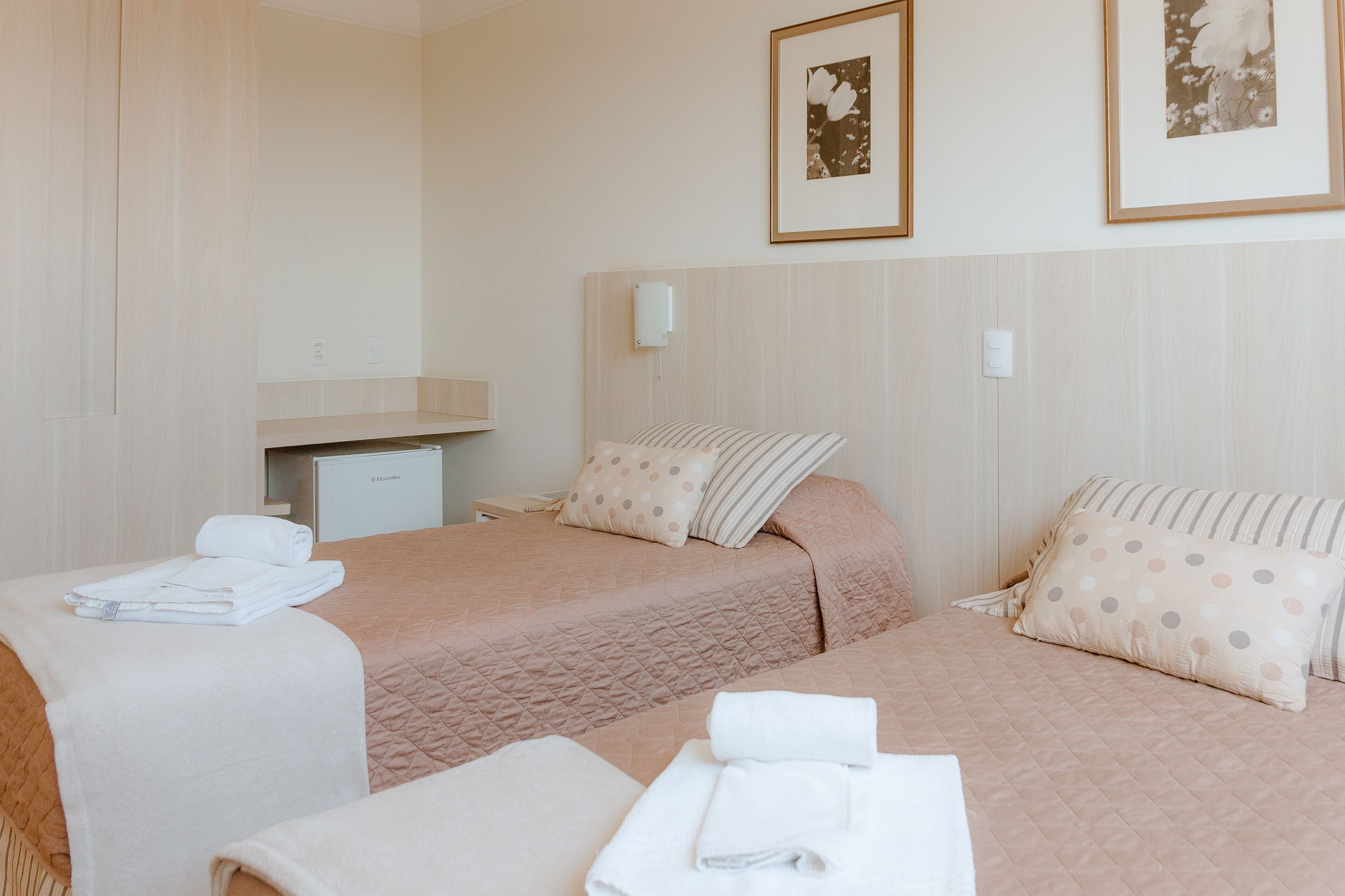 A hotel room with two beds, each with a pink quilted bedspread and matching pillows. There is a small desk with a lamp and a framed picture on the wall above it.