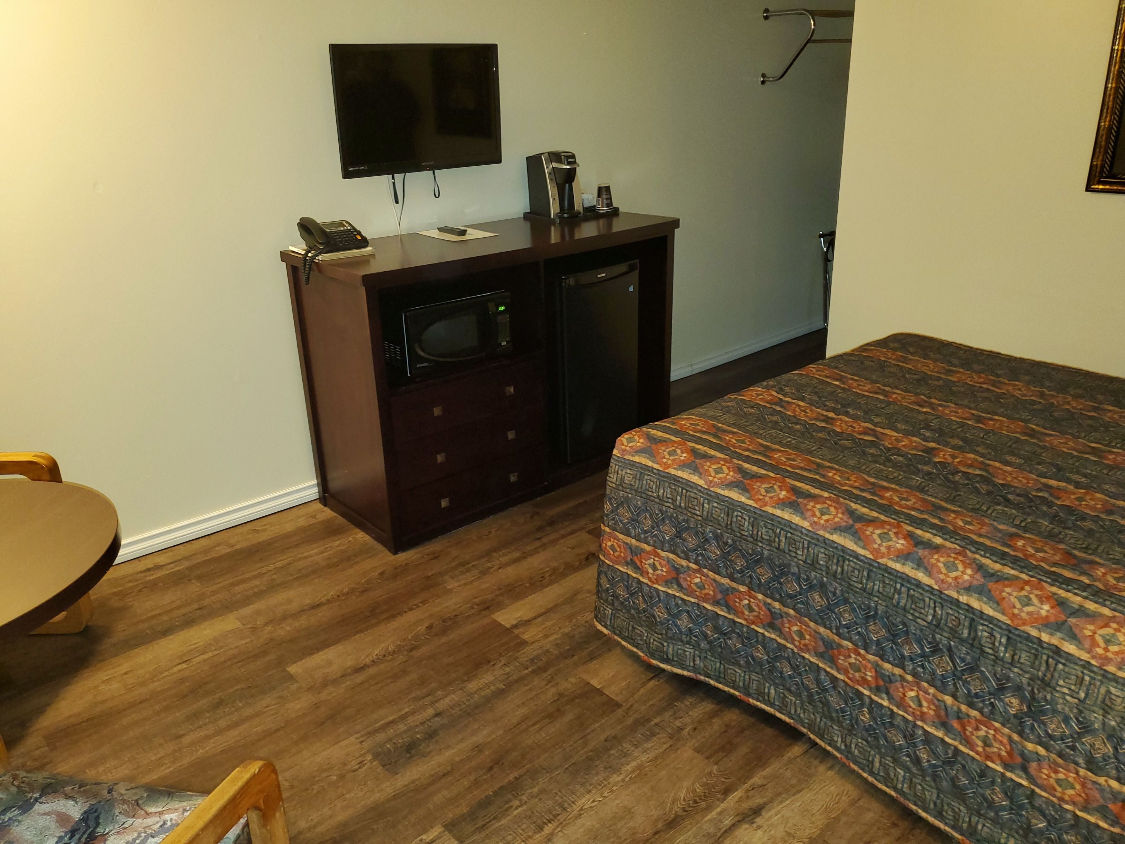 A hotel room with a wooden floor, a bed with a patterned bedspread, a desk with a telephone and a coffee maker, and a TV mounted on the wall.