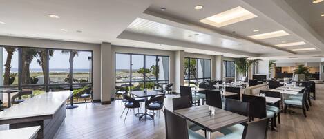 A spacious indoor dining area with multiple tables and chairs, large windows offering a view of the outdoors, and a modern ceiling design.