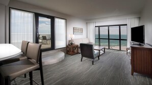 A hotel room with a large sliding glass door to the balcony, a flat-screen TV, a sofa, a dining table, and a small side table. - Island House Hotel Orange Beach - a DoubleTree by Hilton (Orange Beach)
